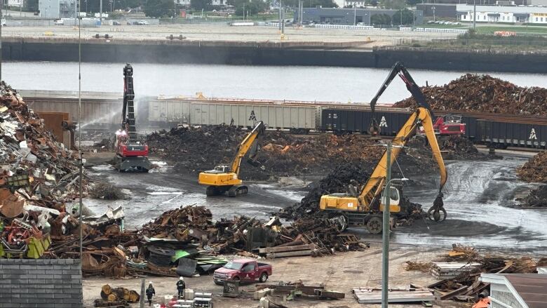 Heavy equipment on a waterfront industrial site with piles of scrap metal.