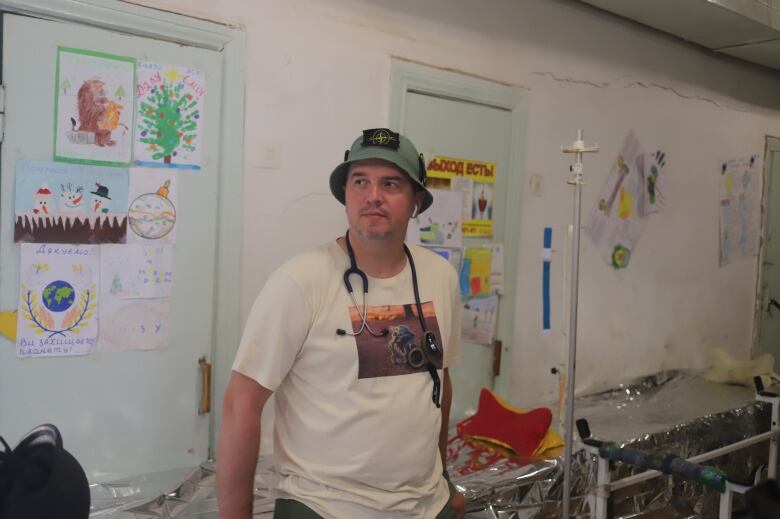 A man wearing a t-shirt, a bucket hat and a stethoscope around his neck stands in a hallway crowded by stretchers and decorated with children's artwork. 