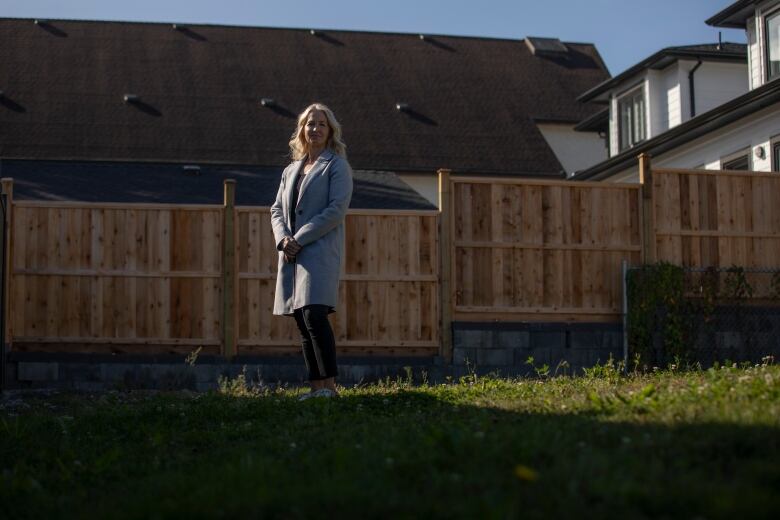 A middle-aged woman with long blond hair and a grey wool coat stands in a barren backyard beside a tall fence.