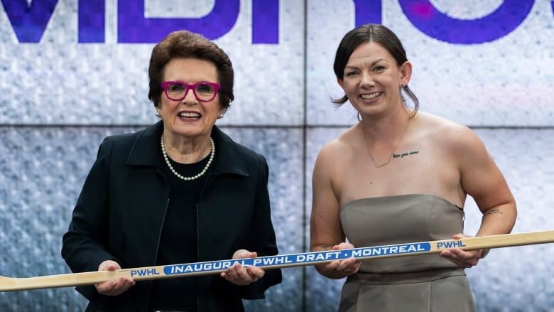 Two women stand on a stage, holding a wooden hockey stick.