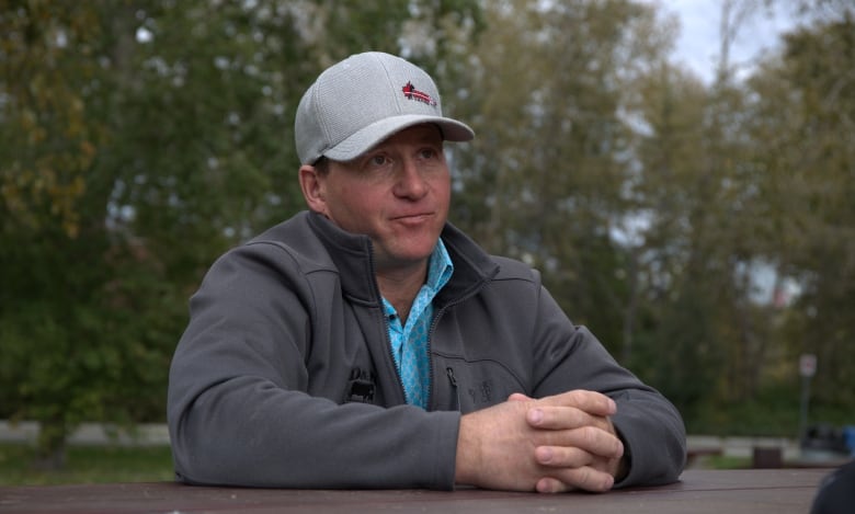 A man wearing a baseball cap leans against a wooden structure with a stand of trees behind him.