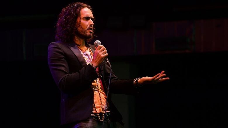 A man in dark jacket holds a microphone as he performs a comedy routine.