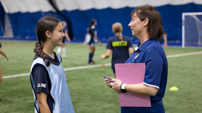 A teenage girl in a light blue soccer vest is shown talking to a female coach with a goal behind them on an indoor field.