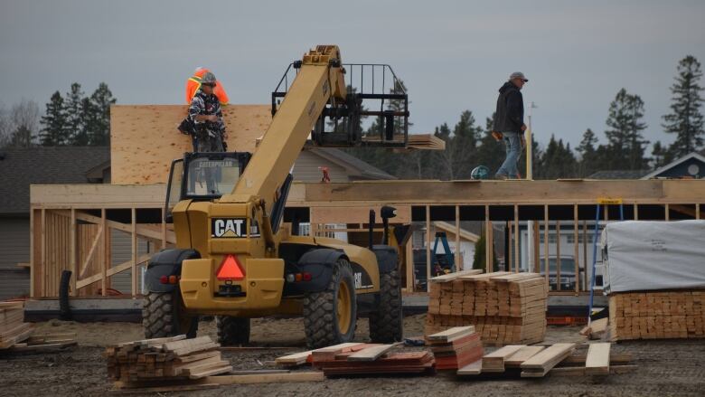 Some men work on a new house, with a machine and a stack of lumber in the foreground