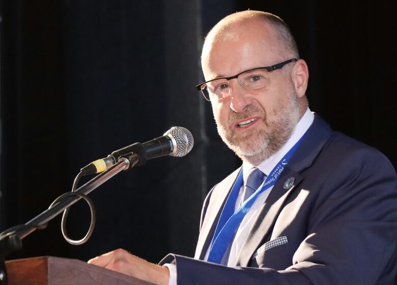 A man with glasses and a blue suit and tie and white shirt speaks at a microphone.