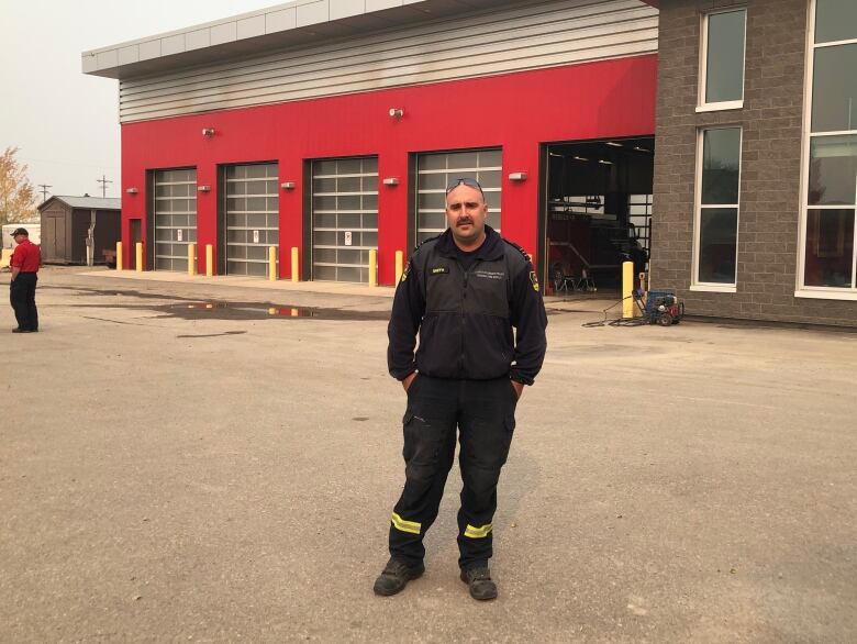 A man in a firefighting uniform stands in the parking lot outside a fire station.