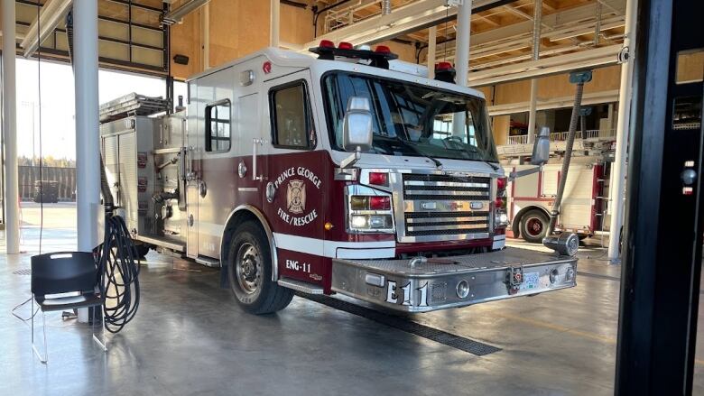 Image shows a red fire truck sitting idle inside a firehall. It says Prince George Fire Rescue on its side. Truck is facing forward and behind it, the garage door is open showing a sunny day outside.