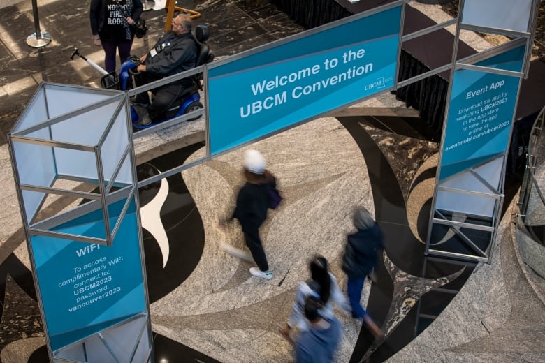 People walk under a banner that says UBCM. Their images are blurred as they are in motion.