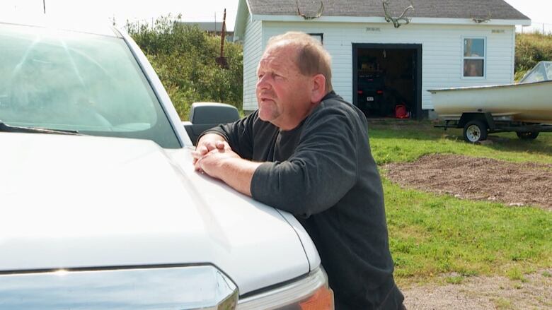 A man wearing a grey sweater leans on a pickup truck with his hands crossed.