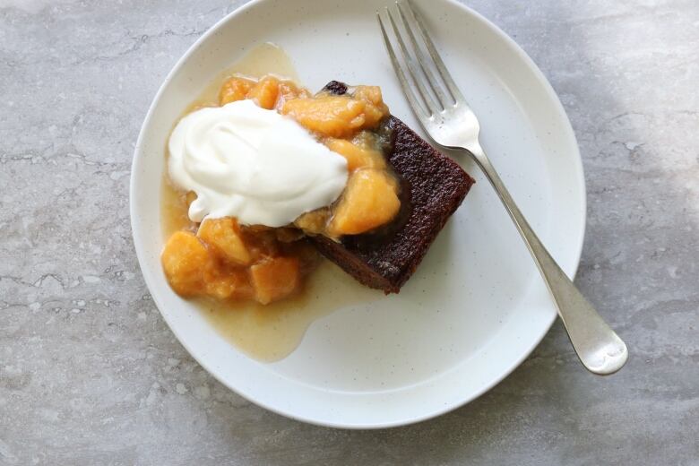 A slice of brown coloured cake topped with peaches and a dollop of whipped cream.  