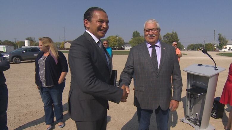 Two men in suits and ties shake hands with each other, in the middle of a parking lot while other people stand around and converse nearby.