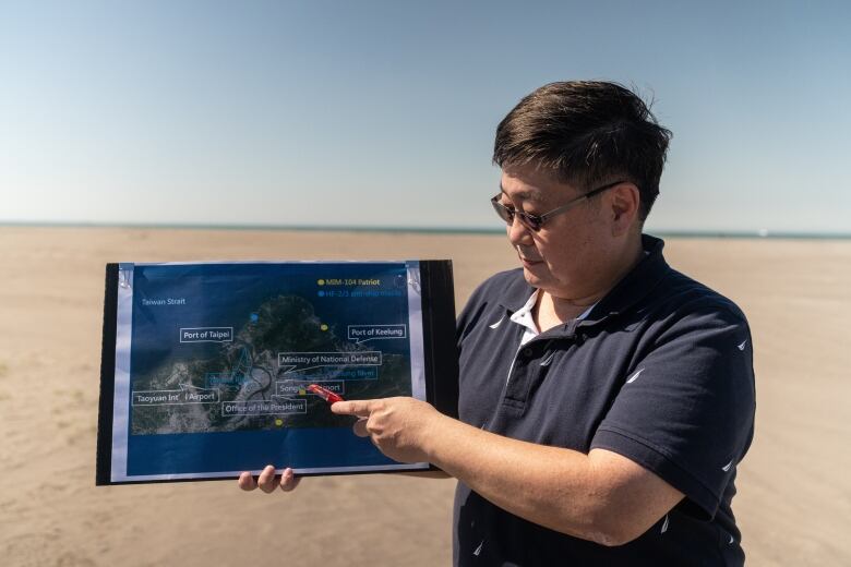 A man wearing glasses stands on a beach and points at a map in his hand.