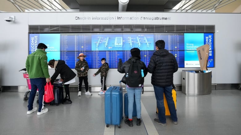 A half dozen people stand in front of a departures and arrivals board.