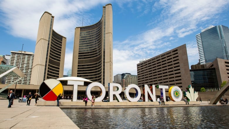 The Toronto sign and City Hall at Nathan Phillips Square on May 11, 2023. Daytime photo. The curved towers of City Hall rise in the background in front of a light-up sign that reads 