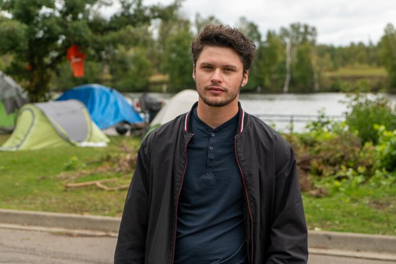 A man stands in a park. Behind him there are tents.