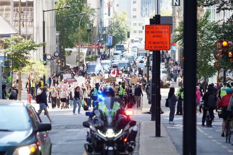 A crowd of people hold signs on a downtown street. A police motorcycle ahead of them.