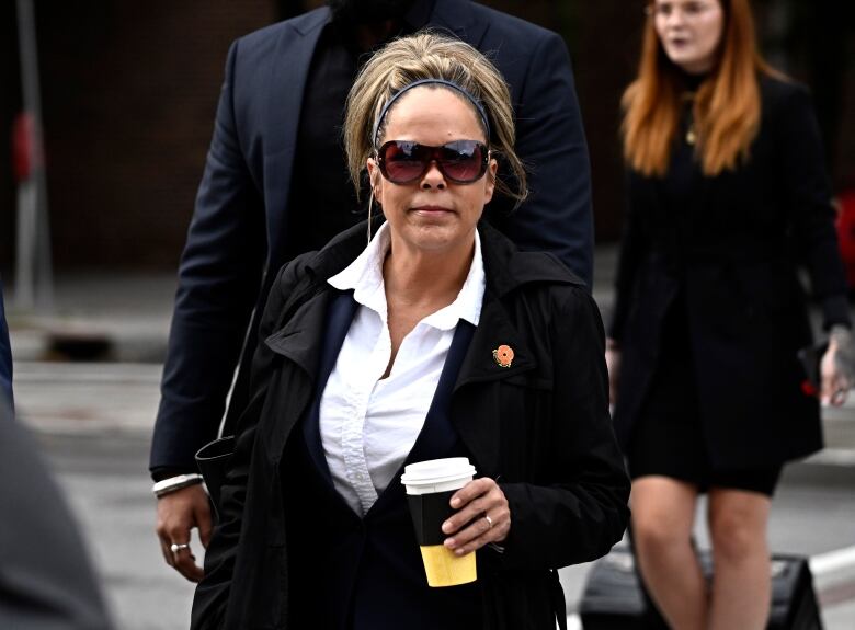 A woman wearing sunglasses carries a disposable coffee cup outside in late summer.