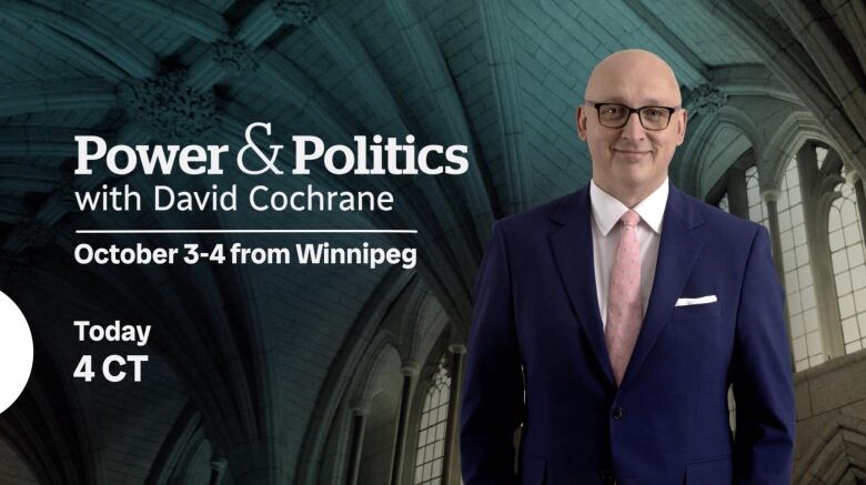 Bald man is wearing black glasses, navy suit and pink tie. Standing beneath arched ceiling of Parliament in Ottawa. 