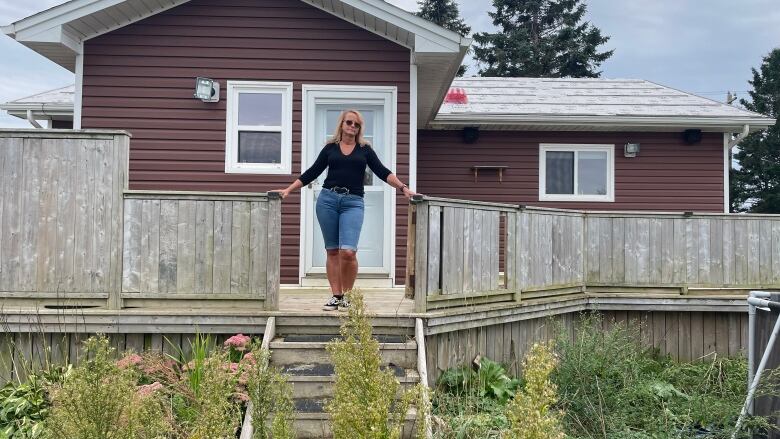 A woman stands at the top of the stairs leading up to a rust-coloured bungalow.