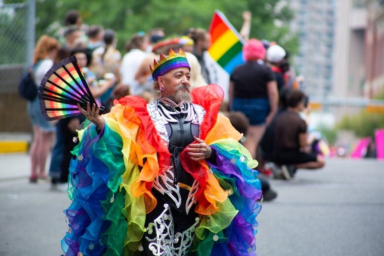 A man holds a rainbow fan while draped in a fluffy rainbow coat. 