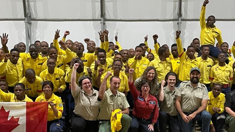 Several South African firefighters clad in yellow jackets pose with 6 B.C. Wildfire Service members. They are cheering and holding up a Canadian flag. 