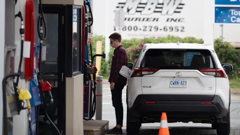 A man with short brown hair and glasses stands at a gas station pump before putting gas in a white car.