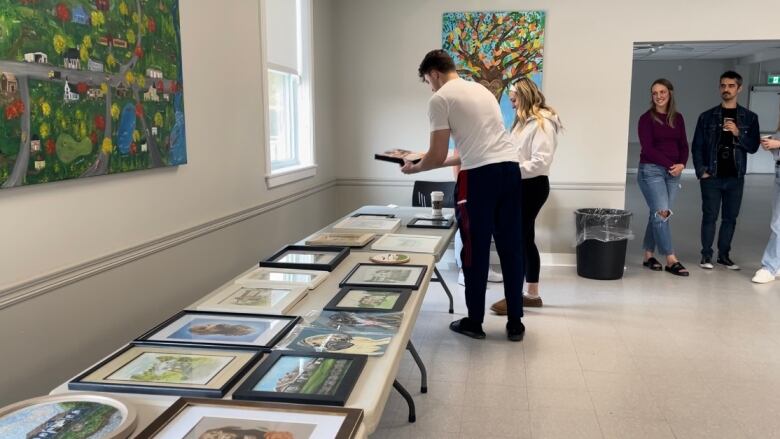 framed paintings and drawings lay out on a long table. A man and woman picked up a drawing and are looking at it. 