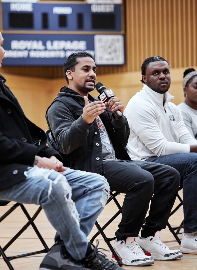 A brown man speaks into a mic while seated on a basketball court, with a Black man to his left.