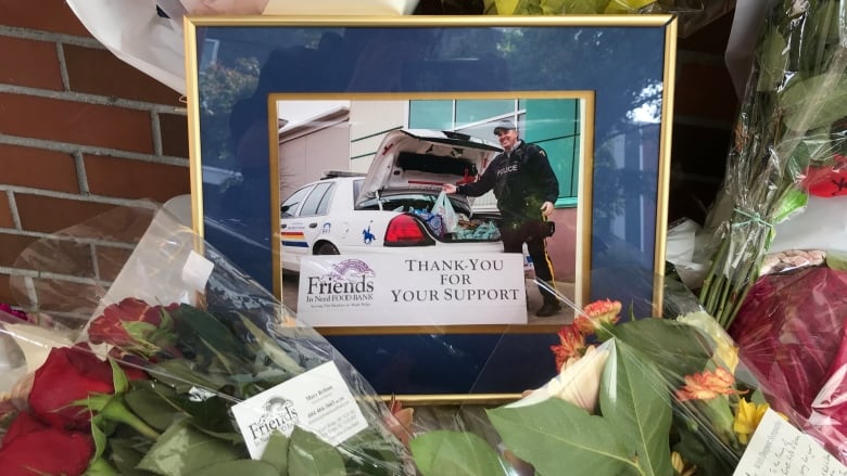  A photos, framed by a navy blue border, is pictured on a while table surrounded by bouquets and unlit candles. The photo shows a man pictured in a black police uniform, taking groceries out of a car's trunk.