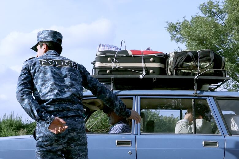 A person in military garb gestures outside a vehicle with suitcases on the roof.