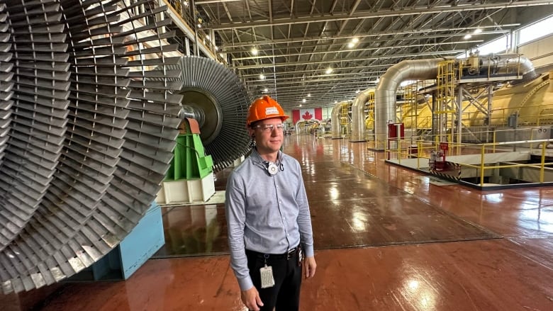 Andrew Galuszka is pictured wearing a hard hat and protective goggles, standing beside a large turbine, with large metal pipes that carry steam to the turbines in the background.   