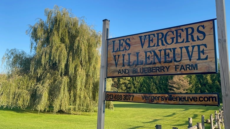 A sign out front of a farm before a well-maintained lawn with trees in the background. 