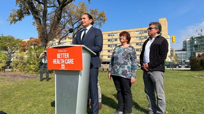 A man stands at a podium in front of two other people. The podium has a sign on it reading 