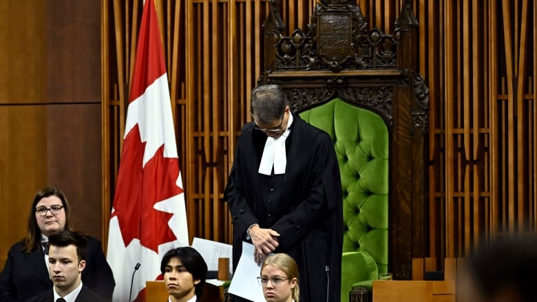 A man in ceremonial robes stands with his head bowed in front of an ornate chair. There is a Canadian flag to his right. 