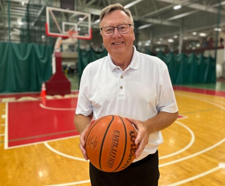 A man wearing a white polo shirt holds a basketball while standing on a court surface. 