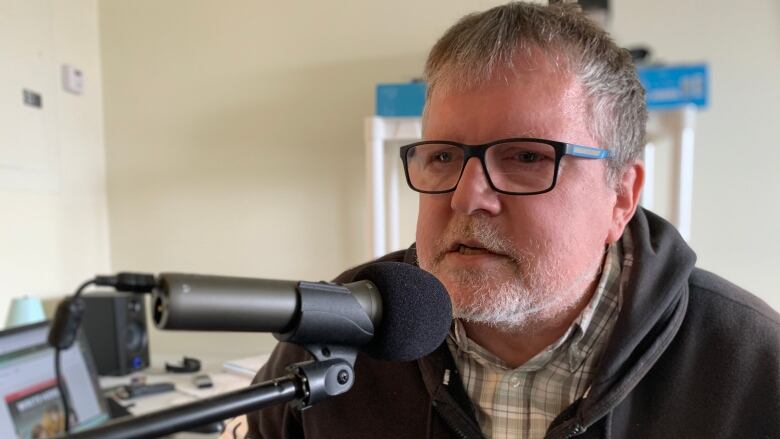 A man with glasses sits in front of a microphone. The man is Darren Surette, who moved in to Affirmative House, an affordable housing development in Dartmouth, Nova Scotia. His $600 a month rent allowed him to go back to school for courses on entrepreneurship and media. He has started his own podcast. 