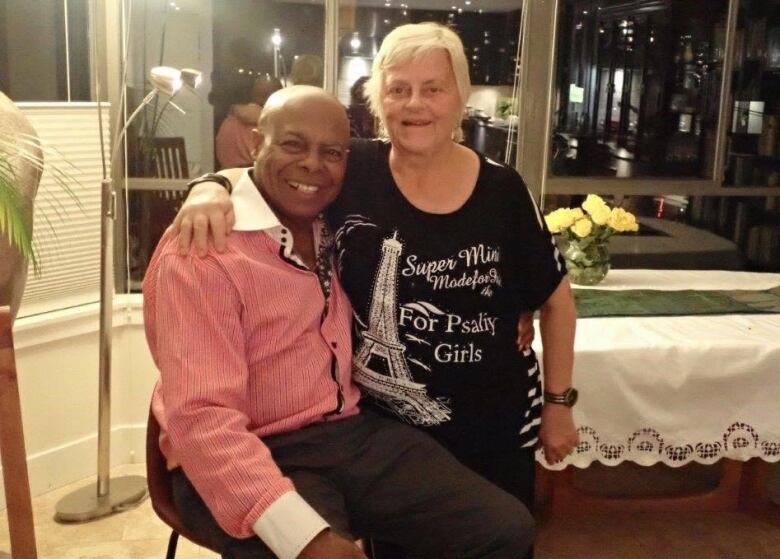 A Black man wearing a pink dress shirt with white cuffs sits on a kitchen chair with his arm around a white woman standing next to him wearing a black t-shirt. They are both smiling.