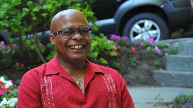 A middle-aged Black man wears a red button up short-sleeved shirt and white pants while sitting on a garden bench holding a wine glass. He has a gold chain around his neck, a gold watch on one wrist and a gold bracelet on the other. He has black aviator sunglasses on and is smiling wide as if possibly in the middle of laughing.