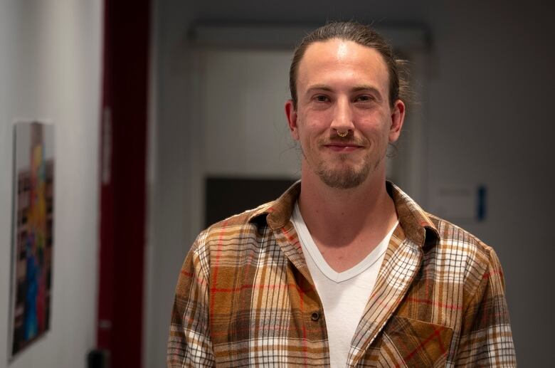 A man with blonde hair in a ponytail, septum piercing and short beard/moustache poses for the camera in a hallway with art hanging on the walls. He's wearing a brown plaid button-up over top of a white t-shirt