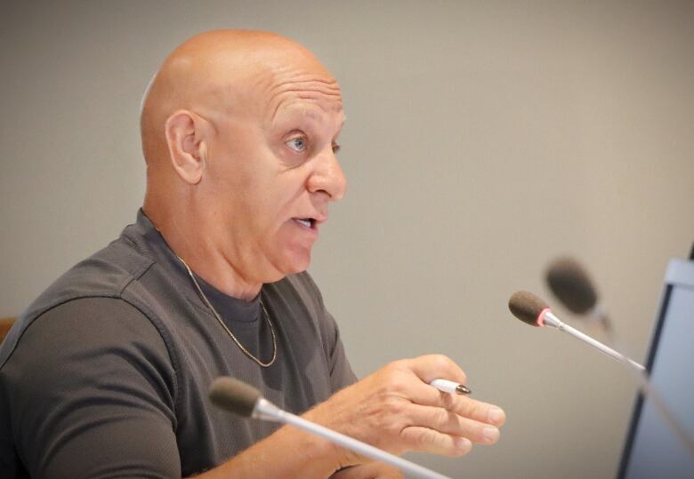 A bald man with a dark grey shirt and gold chain gestures with a pen in his hand while speaking into a microphone.