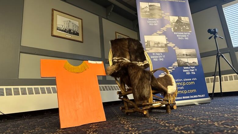 An orange shirt and a chair draped in fur are displayed inside a room, beside a microphone and banner.