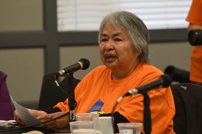 An older woman in an orange t-shirt sits at a table and speaks into a microphone.