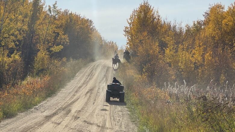 Riders on horseback and one person on an all-terrain vehicle on a dusty road under bright autumn sunshine.