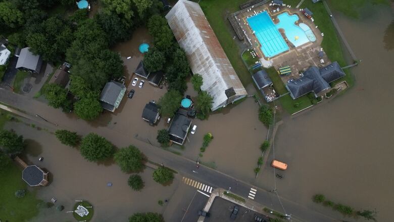 A drone shot shows brown water overflowing into the streets and lawns of a residential area. there's a bright blue swimming pool. 