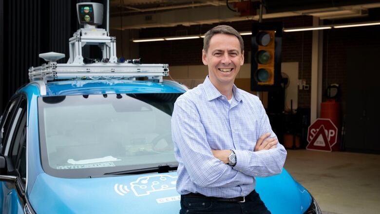 A man stands smiling and arms crossed in front of a parked blue car. The car has cameras and other mechanical things attached to its roof.