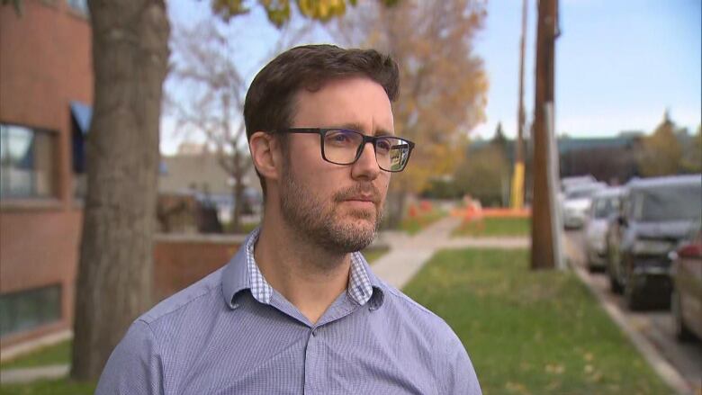 A man looks off camera. He is wearing glasses and standing outdoors. 