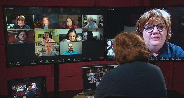 A woman is pictured from behind. In front of her are two screens, one shows a group of people on a Zoom call, and the other shows her face.