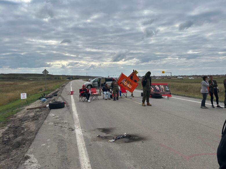 People sit in chairs on a road. A car is parked  behind them.