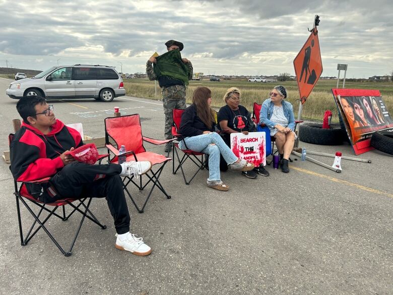 People sit in chairs on a road. Someone holds a sign that says 