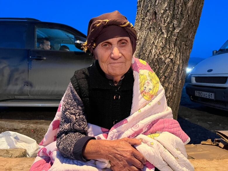 An older woman wrapped in blankets sits on the ground beside a tree.